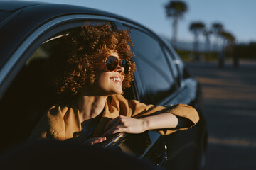 Fototapeta premium Vibrant woman lifestyle outdoors, smiling from a car window, sunglasses on, sunlit breeze, retro film color palette, candid and relaxed moment