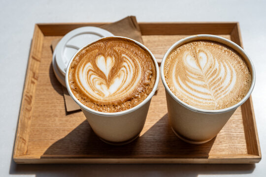 Two takeaway lattes topped with heart-shaped latte art sit on a wooden tray, casting soft shadows and conveying warmth, friendship, and a cozy morning coffee ritual in a minimalist cafe setting.