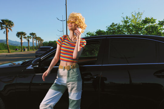 A fashion forward woman in a striped cropped top and patchwork jeans leans against a sleek black car under bright sun, conveying retro lifestyle and street style.