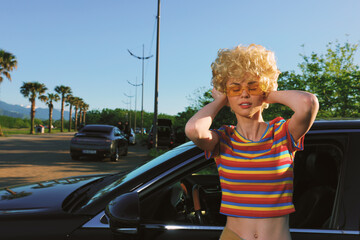 Woman with blonde curly hair wearing a striped rainbow crop top stands beside a parked car on a sunlit street, retro film color mood, casual lifestyle moment, confident pose © SHOTPRIME STUDIO