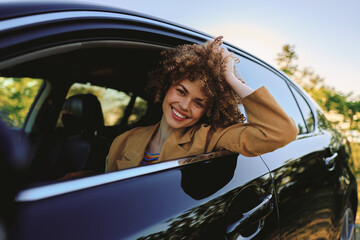 Fototapeta premium A smiling woman leans out of a sleek car window, wearing a casual jacket, as warm sunlight floods the scene. An upbeat lifestyle moment captured outdoors with freedom.