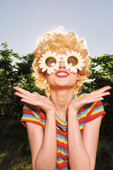 Fototapeta premium A woman in retro colorful dress stands outdoors with flower sunglasses, showcasing lifestyle, portrait, and playful mood in a sunlit park. Bright, cheerful, and fashion-forward, the scene evokes