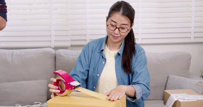 Asian woman small business owner tapes parcel box securely, completing online order packing process, supporting home based e-commerce logistics, preparing shipment for customer delivery