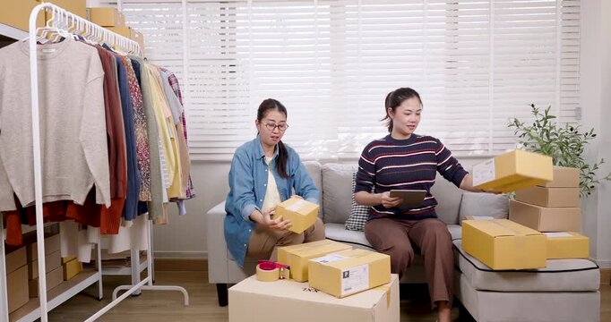 Asian women small business partners working together packing online shopping parcel box, checking customer order list, running daily e-commerce business workflow with teamwork