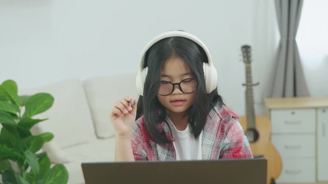 Young girl with glasses and headphones using a laptop computer at home, sitting on a sofa with a guitar nearby, enjoying digital entertainment and learning