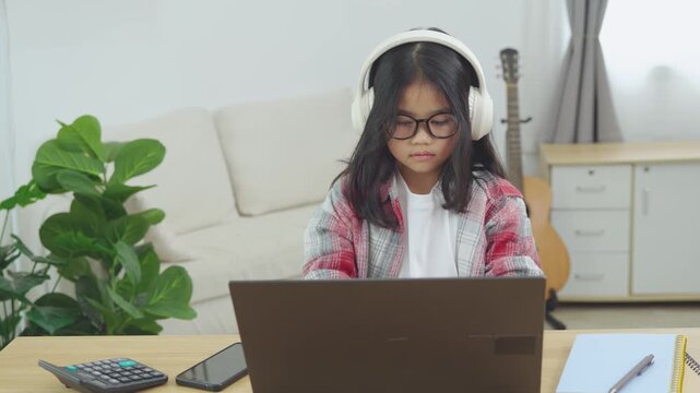 Young girl with long dark hair wearing glasses and headphones using a laptop in a modern living room during daytime, with a guitar and potted plant in the background
