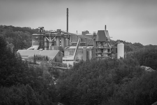 Die Fabrik ruht im grauen Licht des Morgens wie ein schlafender Riese aus Eisen. Zwischen den dunklen Baumwipfeln wirken Rohre, T&uuml;rme und F&ouml;rderb&auml;nder fast wie die Adern einer eigenen kleinen Stadt.