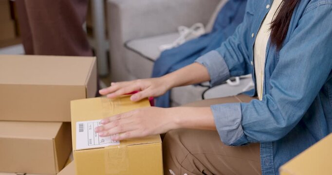 Asian women small business partners sealing online shopping parcel box by hand movement, packing shipment slowly, preparing delivery for customers, managing home warehouse workflow, daily e-commerce