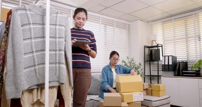 Asian women small business partners manage online shopping parcel box tasks together, checking orders on tablet, preparing e-commerce shipment, coordinating home based online selling workflow