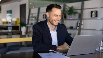 Obraz premium Smiling businessman working on laptop sits in modern office, reviewing documents, reading information with positive business news, engaging in online tasks. Efficiency, successful workflow results