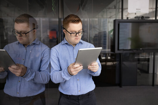Young man with Down syndrome wearing glasses using tablet, working online, standing in modern office, preforming corporate tasks, solve work-related matters remotely, emailing, chatting, strategizing