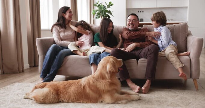 Couple playing with children on sofa, laughing, tickling, having fun, enjoying playtime, spending weekend leisure together in living room at home with golden retriever dog. Large family bonds and love