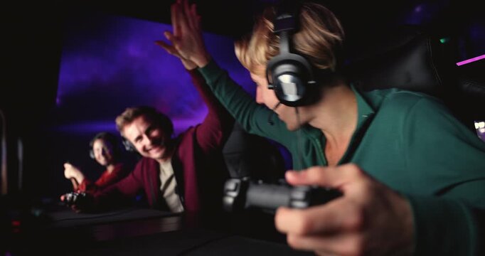 Young adult male gamer giving high five to teammate, celebrating video game victory, playing videogame with controller in cybercafe club. Competitive esports activity, online entertainment, hobby