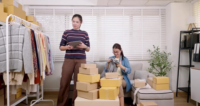 Asian women small business partners managing online store tasks, checking parcel box orders, coordinating packing workflow, organizing home warehouse inventory, supporting daily e-commerce operation