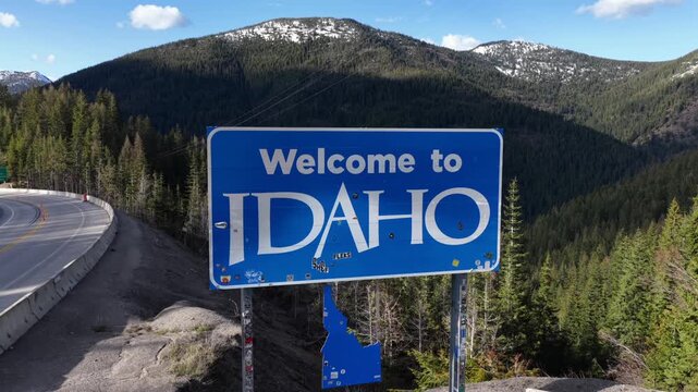Welcome to Idaho blue road sign on mountain pass highway surrounded by green pine forest and snow capped peaks under clear sky during sunny spring daytime in American Pacific Northwest. Aerial view.