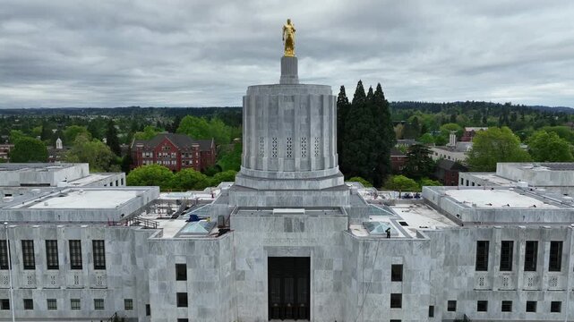 Oregon State Capitol in Salem with gold Pioneer statue atop Art Deco dome with worker on roof and green trees under overcast sky during spring daytime in American Pacific Northwest. Aerial rising view