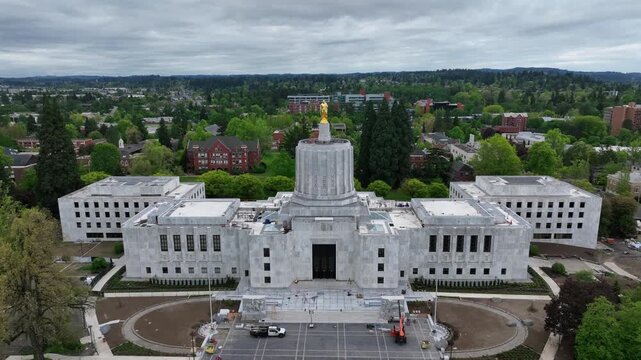 Oregon State Capitol building in Salem with white marble facade and Art Deco dome with golden statue surrounded by lush green trees under overcast sky during spring in United States. Aerial rise wide