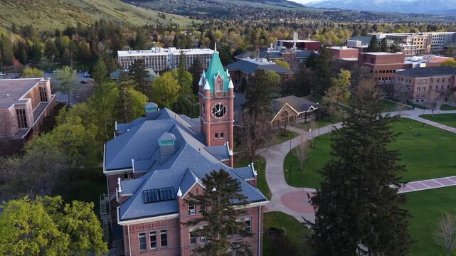 University of Montana campus in Missoula with historic Main Hall and clock tower at sunrise with snowy mountain peaks in background and green trees during spring in United States. Aerial wide shot.