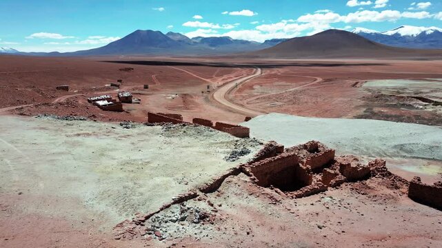 Forward flight over ancient atacame&ntilde;o stone corral ruins on red desert plateau with winding dirt road and snow-capped volcanic peaks under blue sky. Putana, Chile