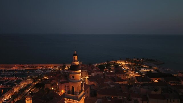 Basilique Saint-Michel Archange bell tower illuminated against deep blue night sky, Menton, France, aerial