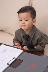 Naklejka premium A 2-year-old toddler boy sitting on the sofa looking to the side with clipboard in front