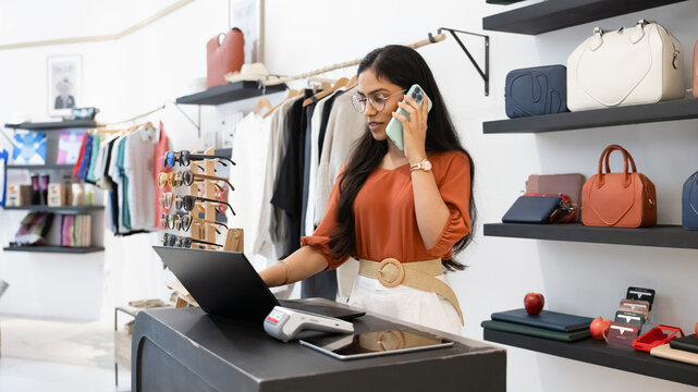 Indian woman stands at checkout counter of modern boutique, having business conversation on phone, working on laptop, check stock, managing online orders, handling customer service or logistics tasks