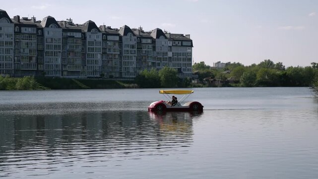 Silhouette of two people driving a pedal boat that looks like a car. Pedalo on water with urban background. Summer active pastime.