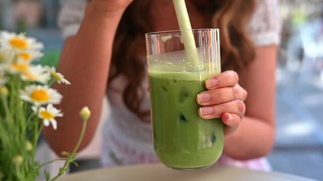 Woman holding glass of matcha latte with ice cubes and drinking with straw outdoors.