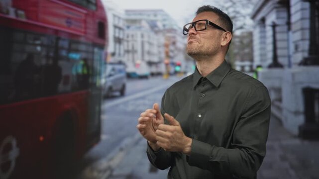 Man with glasses clapping hands beside red double decker bus on street while smiling and giving thumbs up; approval.