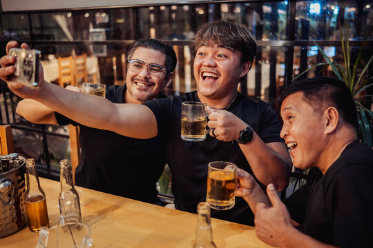 Three friends happily video chatting for their friend in another country at a bar while holding cold beer mugs, enjoying nightlife, bonding, and celebrating friendship together.
