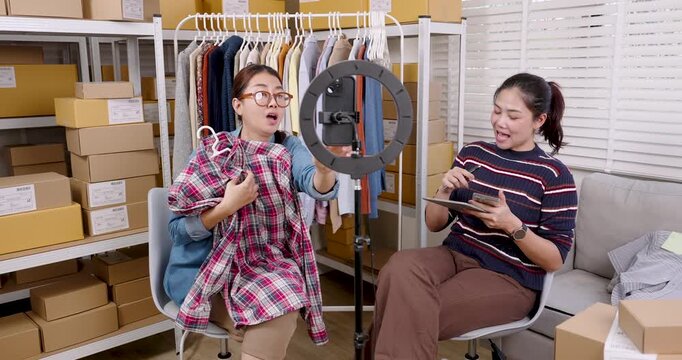 Asian women partners presenting clothing item to camera during live selling, discussing product features, checking online order list, managing e-commerce workflow inside home warehouse studio