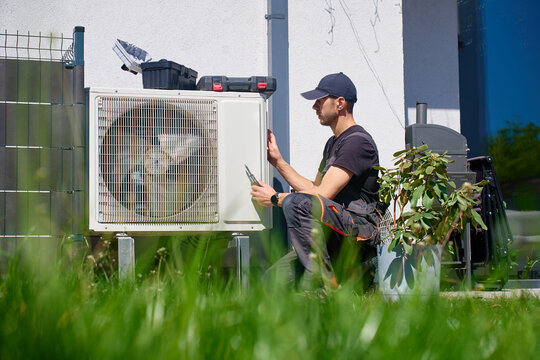 HVAC technician services an outdoor unit near a house wall, holding a tool beside the grille. Concept of home heating, cooling and maintenance.