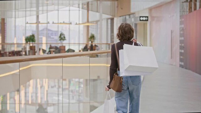 Rear view slowmo shot of female customer walking through shopping mall with modern glass interior carrying bags on shoulder