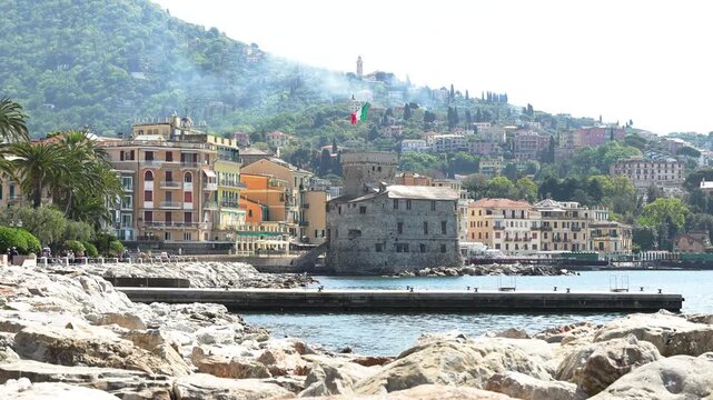 Wide angle view of Rapallo Castle and vibrant Ligurian architecture by the sea under bright daylight, with rocky shoreline, lush green hills, and clear blue water. No people are visible.