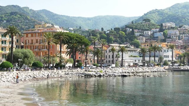 Wide shot of Rapallo&rsquo;s picturesque waterfront, palm-lined promenade, pastel villas, calm Ligurian Sea, and lush hills. Clear daylight, vibrant architecture, gentle water, serene Mediterranean mood.