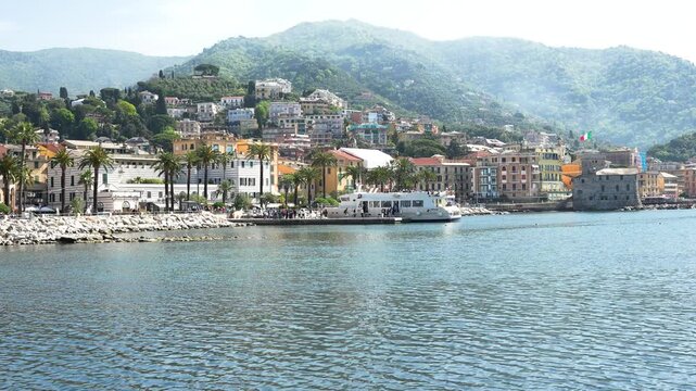 Wide angle shot of Rapallo's picturesque harbor, lined with vibrant architecture, palm trees and calm blue waters, backdropped by lush green hills under soft natural daylight. (198 characters)
