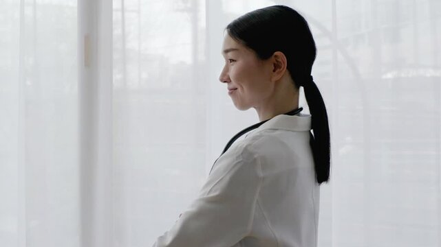 Asian Female Doctor in White Coat Smiling with Arms Crossed by Bright Window