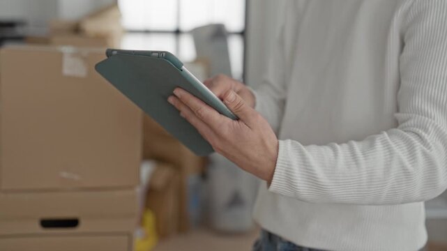 Man holding tablet and checking stacked cardboard moving boxes and parcels in building; focused inventory management.