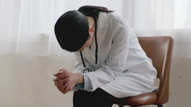 Asian Female Doctor in White Coat Sitting with Head Down, Exhausted