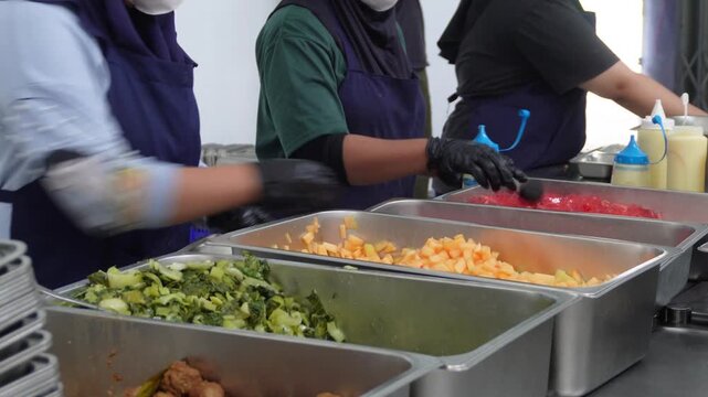 Kitchen staff wearing black gloves preparing food portions from stainless steel buffet trays, commercial catering service, restaurant meal prep, and food assembly concept.