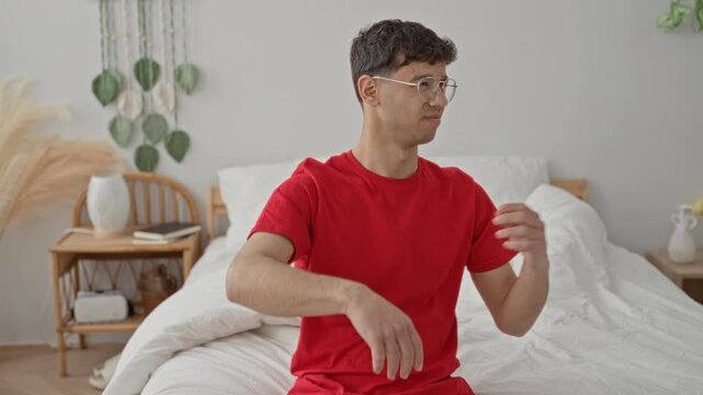 Young man in red shirt sits on bed and wipes his face with his forearm while squinting and swatting at his face inside a building; discomfort.