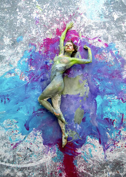 A nude woman lies on a large abstract canvas, her body covered in green and gray paint. Surrounding her are vivid blue, purple, and pink splashes, merging figure and painting into one expressive form
