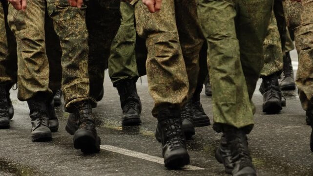 Close-up view of marching soldiers in camouflage uniforms and black boots on a wet city street, showing disciplined formation and synchronized steps in a coordinated military drill