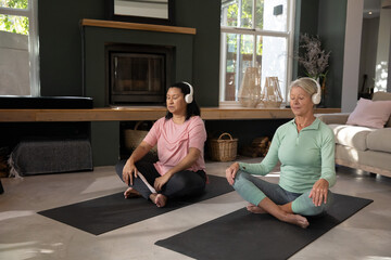Fototapeta premium Mature, senior women friends meditating on black yoga mats at home in activewear wearing headphones