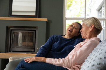 Fototapeta premium Mature female friends laughing and chatting on sofa in living room near fireplace