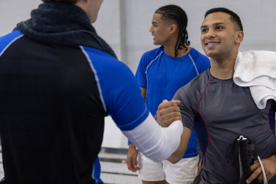 Diverse male players in jerseys clasping hands, smiling at gym with prosthetic blade and towel