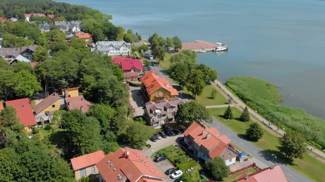 An aerial shot captures the picturesque charm of a small coastal town in Lithuania, likely Neringa or the Curonian Spit area. The scene features traditional, red roofed buildings nestled among lush gr