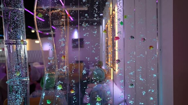 Decorative bubble wall panel features rising air bubbles and colorful toy fish floating within illuminated vertical glass chambers of water.