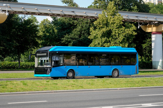 A blue electric bus travels along a city street past a green park and a viaduct, side view