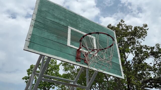 Old outdoor basketball hoop on a green wooden backboard under a clear blue sky. Tilt up motion